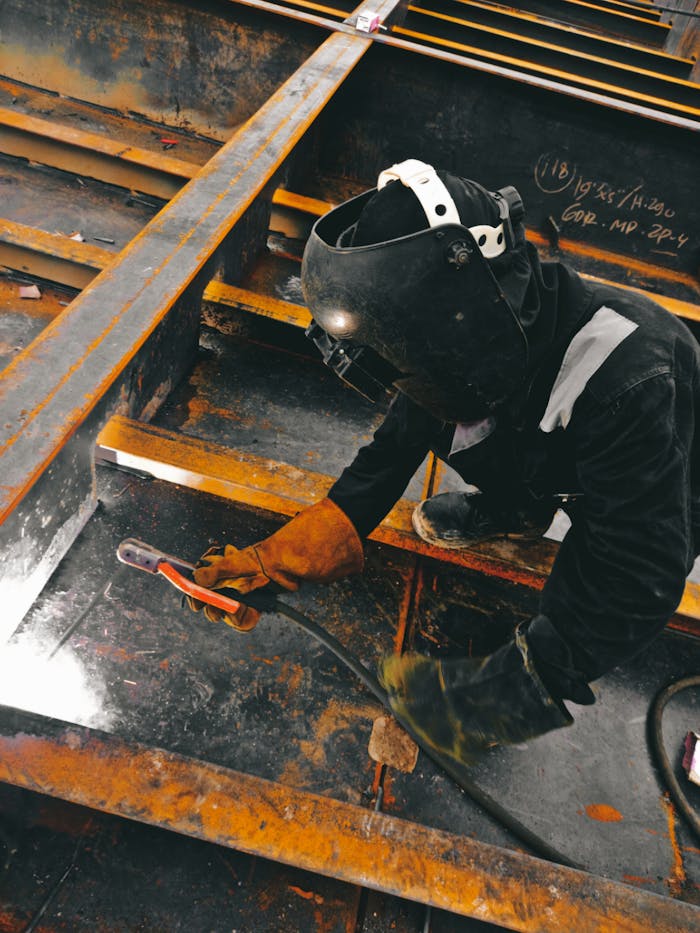 Home A welder in action wearing protective gear and welding helmet in an industrial setting.