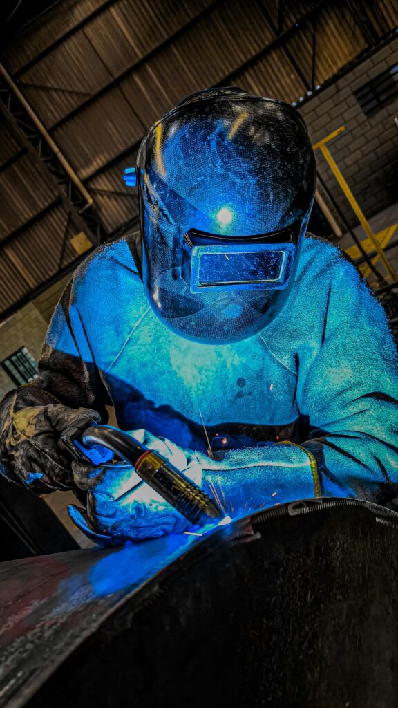A welder in full protective gear working with a bright blue arc in a Brazilian industrial setting.